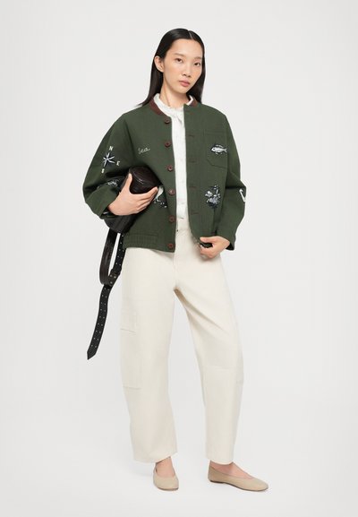 Chaqueta bomber de algodón verde con diseños bordados, cierre de botones, una camisa blanca debajo, pantalones anchos crema y bailarinas beige.