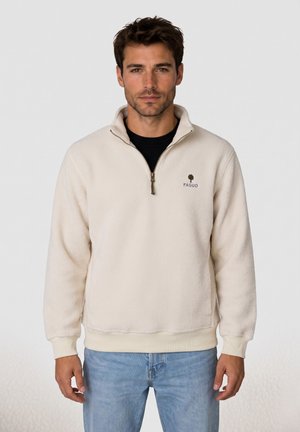 Man draagt een crèmekleurige fleece trui met een kwart rits en "FAGUO" logo, gecombineerd met lichtblauwe spijkerbroek, staand tegen een effen achtergrond.