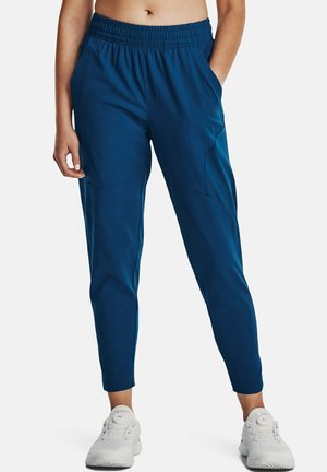 Person trägt blaue, verjüngte Sporthosen und weiße Laufschuhe, steht mit einer Hand in der Tasche vor einem einfarbigen Hintergrund.