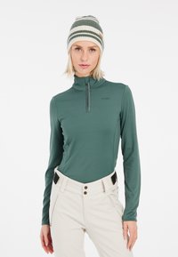 Maglia verde a maniche lunghe con zip realizzata in tessuto morbido, abbinata a pantaloni chiari e un berretto in maglia a righe multiple.