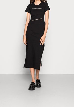 Donna che indossa un abito midi nero con maniche corte, due zip argentate inclinate sul davanti e mocassini chunky neri.