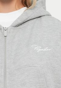 Siva zip-up dukserica od mekog materijala s prednjim džepom i suptilnom bijelom vezom "Pegador" na prsima.