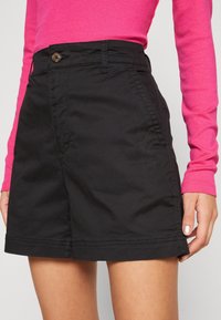 Pantaloni corti neri in cotone con una texture liscia, dotati di chiusura con bottone, tasche laterali e taglio dritto fino all'orlo.
