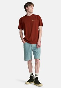 Camiseta de algodón color óxido con un logo sutil, combinada con pantalones cortos de mezclilla azul claro. Zapatillas altas negras y calcetines a rayas completan el look.