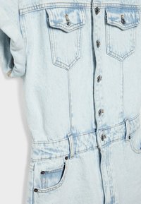 Combinaison en denim bleu clair à manches courtes, fermeture à boutons sur le devant, deux poches poitrine et taille cintrée avec ceinture. Présente une texture délavée.