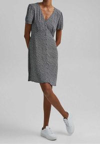Kurzärmliges, V-Ausschnitt Kleid in Schwarz mit einem weißen Blumenmuster, das eine lockere Passform und Raffung im Bereich der Brust aufweist. Getragen mit weißen Sneakers.