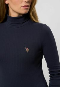 Femeie îmbrăcată cu un pulover din tricot cu guler înalt de culoare bleumarin, având un mic logo brodat cu jucător de polo pe piept, așezată spre stânga, pe un fundal simplu.