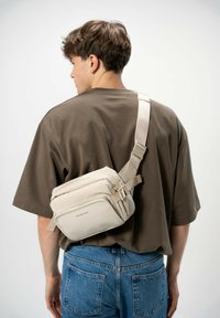Borsa a tracolla beige realizzata in materiale morbido, con più scomparti con zip, tracolla regolabile e design semplice. Indossata sopra una camicia marrone.