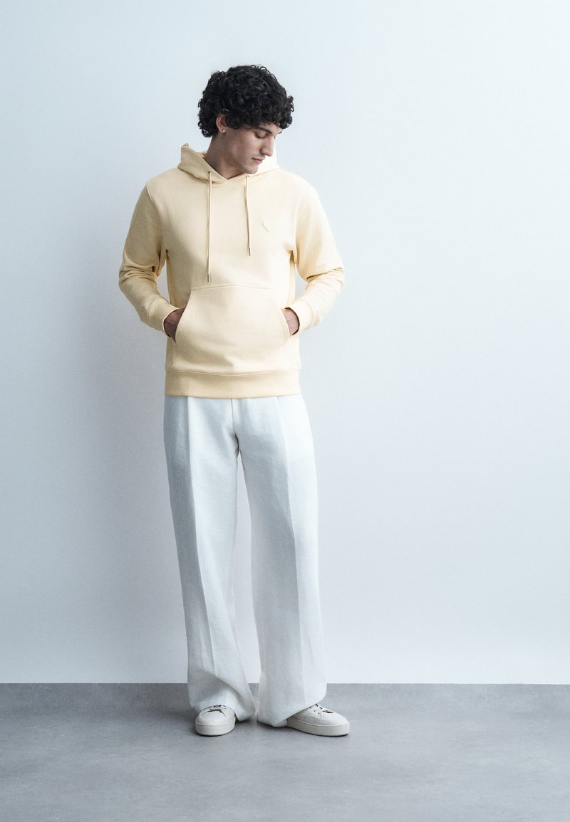 Young man with curly hair wearing a beige hoodie, white wide-leg pants, and white sneakers standing against a plain light background.
