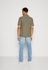 Camisa de manga corta de color verde oliva con una textura en la tela, combinada con jeans de color azul claro. El conjunto tiene un corte relajado y un diseño informal.