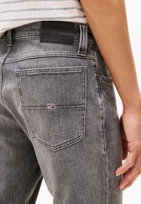 Grå denim jeans med en svart lærbeltelapp merket "TOMMY JEANS." Baklommen har et lite logo i rødt, hvitt og blått.