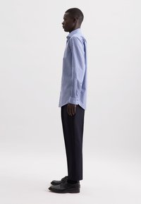 Uomo in profilo, con una camicia a righe blu e bianche, pantaloni scuri e scarpe eleganti nere, su uno sfondo neutro.