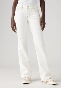 Hvite vide jeans med lys søm, høy midje og frontlommer, iført med beige sneakers. Ensfarget bakgrunn, fokusert på buksene.