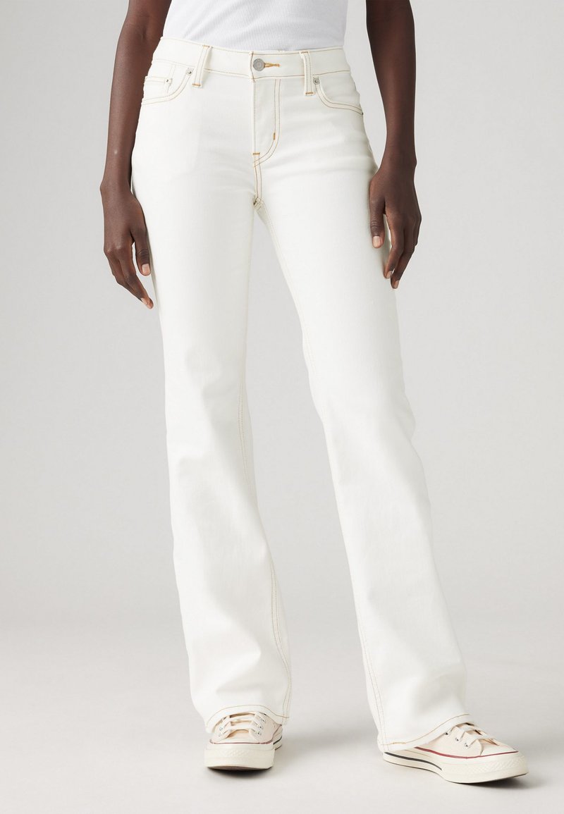Jean flare blanc avec des coutures claires, taille haute et poches avant, portés avec des baskets beiges. Fond uni, axé sur le pantalon.