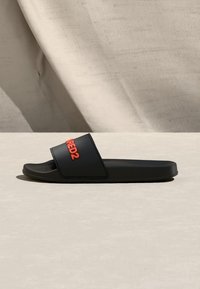Μαύρο σανδάλι slide με φαρδιά λουρί που φέρει κόκκινο κείμενο "RED2", εμφανιζόμενο σε πλευρικό προφίλ πάνω σε ανοιχτό μπεζ επιφάνεια και φόντο υφάσματος.