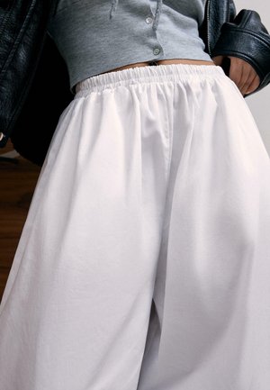 Person trägt lockere weiße Hose mit elastischem Bund, kurzes graues Oberteil mit Knöpfen und schwarze Lederjacke über der Schulter.