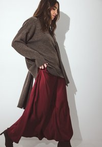 Brauner, übergroßer Strickpullover, kombiniert mit einem fließenden burgunderfarbenen Satin-Maxirock. Glatte Textur, lässige Passform und sichtbare Layering-Designelemente.