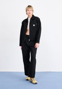 Jachetă neagră cu fermoar, buzunar frontal și patch cu logo, asortată cu pantaloni negri și adidași galbeni. Fundal deschis.