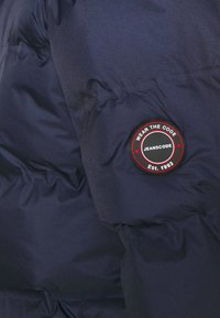 Giacca puffer blu marina con tessuto testurizzato, design trapuntato e una patch con logo circolare che riporta la scritta "Wear the Code, Jeanscode, Est. 1993."