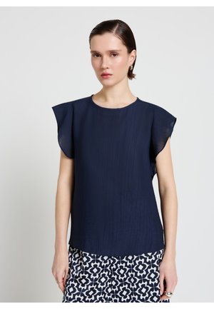 Frau mit kurzen dunklen Haaren, die eine dunkelblaue ärmellose Bluse und einen gemusterten schwarz-weißen Rock trägt, steht vor einem schlichten hellen Hintergrund.