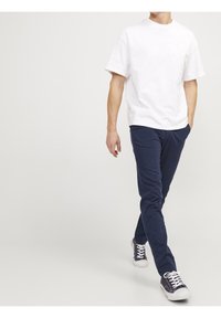 Homme marchant portant un t-shirt blanc uni, un pantalon bleu marine et des baskets noires avec des lacets blancs sur un fond clair.