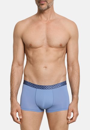 Mężczyzna bez koszulki, ubrany w jasnoniebieskie bokserki z ciemnoniebieskim, wzorzystym pasem, stojący na jasnym, jednolitym tle.