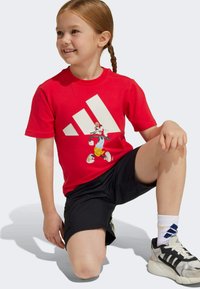 Červené bavlněné tričko s grafikou postavy na přední straně, v kombinaci s černými šortkami a sportovními botami, umístěné na jednobarevném pozadí.