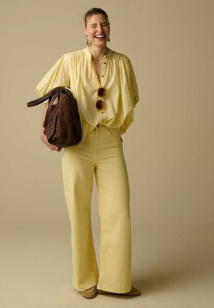 Femme riant les yeux fermés portant un chemisier ample jaune, un pantalon jaune à jambes larges, tenant un sac imprimé léopard et des lunettes de soleil posées sur le chemisier.