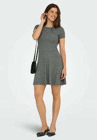 Femme souriante, portant une robe grise à manches courtes avec des chaussures à talons noires et un petit sac à bandoulière noir, posant avec la main près des cheveux.
