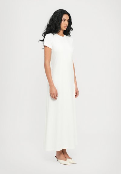 Abito maxi bianco con maniche corte, silhouette dritta e texture del tessuto liscia. Indossato con mule a punta e capelli leggermente ondulati.