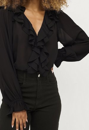 Kvinde iført sort bluse med flæset V-udskæring og lange puffede ærmer kombineret med sorte bukser med høj talje, hånd på hoften.