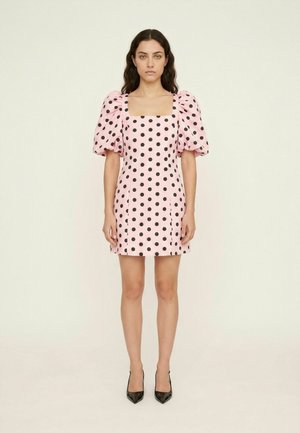 Femme debout, portant une mini-robe rose à pois noirs, manches courtes bouffantes, encolure carrée et talons noirs pointus.