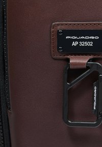 Borsa in pelle marrone con clip e targhetta in metallo nero, caratterizzata da una superficie texturizzata e dettagli cuciti. La targhetta riporta il nome del marchio e il numero del modello.