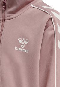 Rosa zip-up jacka gjord av mjukt tyg. Har en vit logotyp och detaljer, med tonala ränder längs axlarna och strukturerat material.