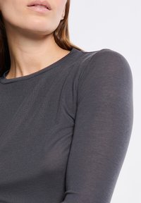 Primo piano di una donna che indossa un top aderente grigio scuro a maniche lunghe, che mette in mostra il décolleté, la spalla e parte del suo viso e dei suoi capelli.