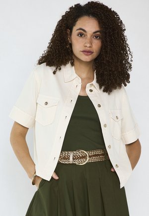 Donna con capelli ricci che indossa una camicia bianca a maniche corte con bottoni sopra una maglietta verde oliva e una gonna abbinata con cintura intrecciata.