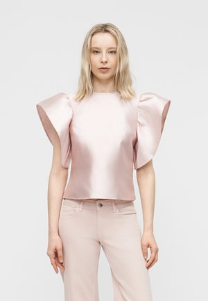 Kobieta o prostych blond włosach, ubrana w blado różową satynową bluzkę z dużymi falbankami na rękawach oraz dopasowane jasnoróżowe spodnie na jednolitym tle.