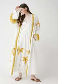 Valkoinen pitkä mekko, jossa on keltaista palmunlehtikirjontaa ja fringe-reunus. Päällä avonainen valkoinen liivi mekko. Mallilla on jalassa matalat sandaalit.