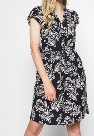 Femme portant une robe noire à manches courtes avec un imprimé floral blanc, des boutons sur le devant et une ceinture nouée à la taille.