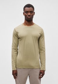 Ilgų rankovių marškiniai alyvuogių spalvos, pagaminti iš švelnios medvilnės, su apvaliu iškirpimu ir pritvirtinta siluetu. Lygus audinys, be matomų raštų.