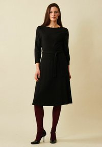 Musta polvimittainen mekko, jossa on istuva yläosa, kolmannesmittaiset hihat ja sidottu vyötäröyksityiskohta. Sujuva kangas ja valuväri.