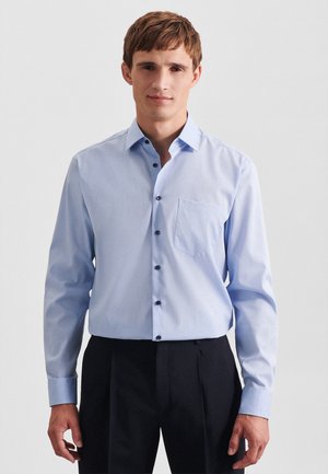 Jonge man die een lichtblauw overhemd met lange mouwen en zwarte broek draagt, staand tegen een effen lichte achtergrond.