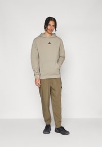 Beige hoodie met kangoeroezak en zwart logo; gecombineerd met olijfgroene cargobroek; zwarte sportieve schoenen maken de outfit compleet.