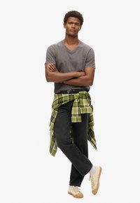 Camiseta gris de cuello en V, jeans negros, camisa a cuadros verde atada alrededor de la cintura, zapatillas color crema, de pie con los brazos cruzados, fondo neutro.