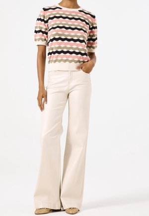 Henkilö, joka käyttää lyhythihaista villapaitaa, jossa on aaltoilevia raitoja vaaleanpunaisena, vihreänä, mustana ja beigenä, leveälahkeisia kerma-colored housuja ja käärmekuvioisia kenkiä.