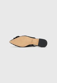 Chaussure tissée noire avec une semelle plate et une semelle intérieure beige lisse. Présente un bout pointu et un motif de crampons minimal sur un talon en caoutchouc noir.