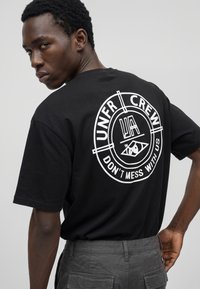 Mann trägt schwarzes T-Shirt mit weißem rundem Logo und Text „UNFR CREW DON'T MESS WITH US“ auf dem Rücken, schaut über die Schulter zurück.