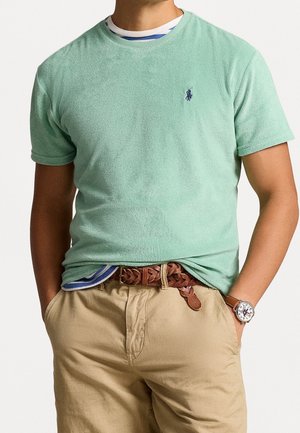 Man draagt een lichtgroen shirt met korte mouwen, een gestreept ondershirt, beige broek met een gevlochten bruine leren riem en een polshorloge met een witte wijzerplaat.