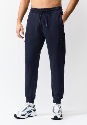 Muž v tmavě modrých teplákových kalhotách s postranními kapsami a bílými teniskami Nike, stojící před bílým pozadím.