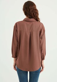 Camicia marrone a maniche lunghe con bottone e colletto, caratterizzata da una plissettatura posteriore e orlo stondato. Tessuto liscio, vestibilità comoda.